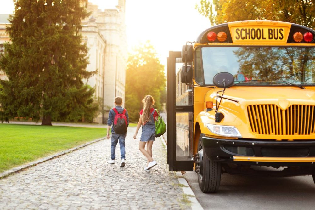 A yellow school bus with flashing lights, stopped on a road with students boarding, highlighting the importance of school bus safety for drivers.