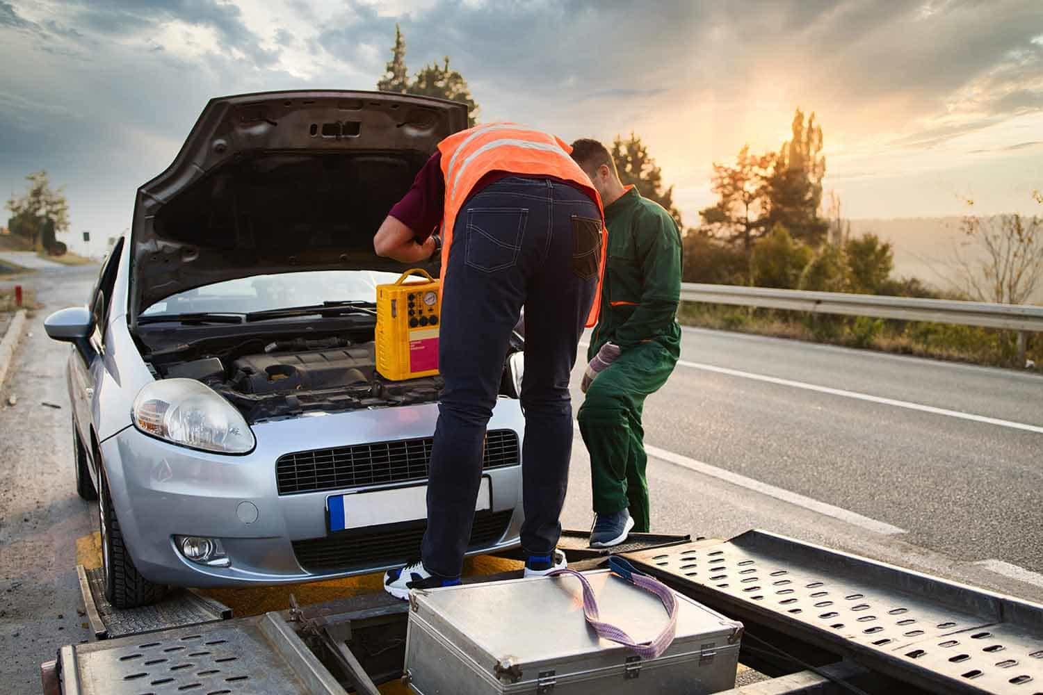 A stranded car on the side of the road with a roadside assistance service vehicle helping to change a flat tire.