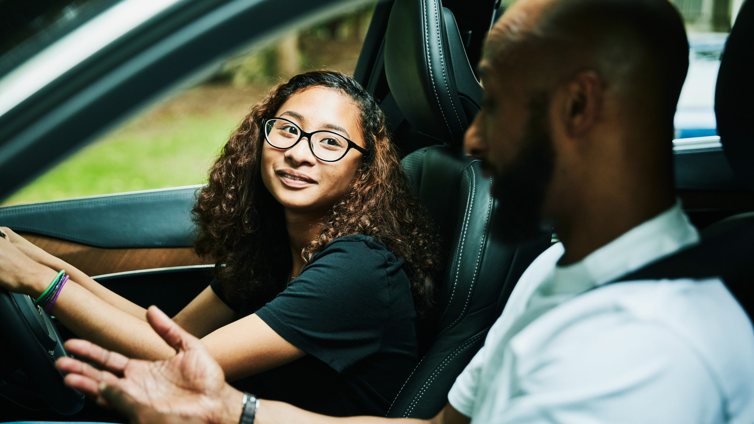 A parent teaching their teenager to drive in a car, with both smiling and focused on the road ahead.